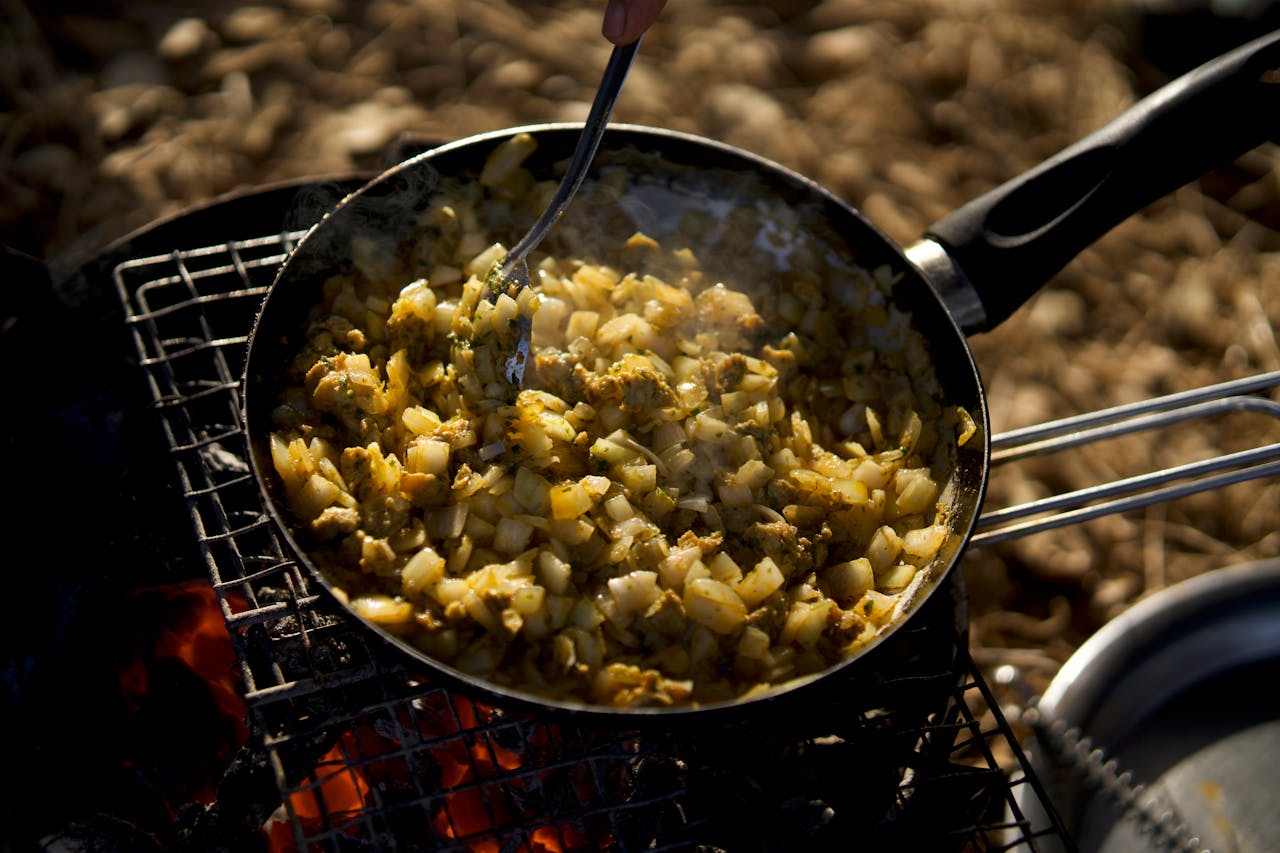 Close-up of a skillet with vegetables being cooked outdoors over an open flame.